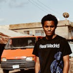 A young man poses outdoors in casual attire beside an orange van.