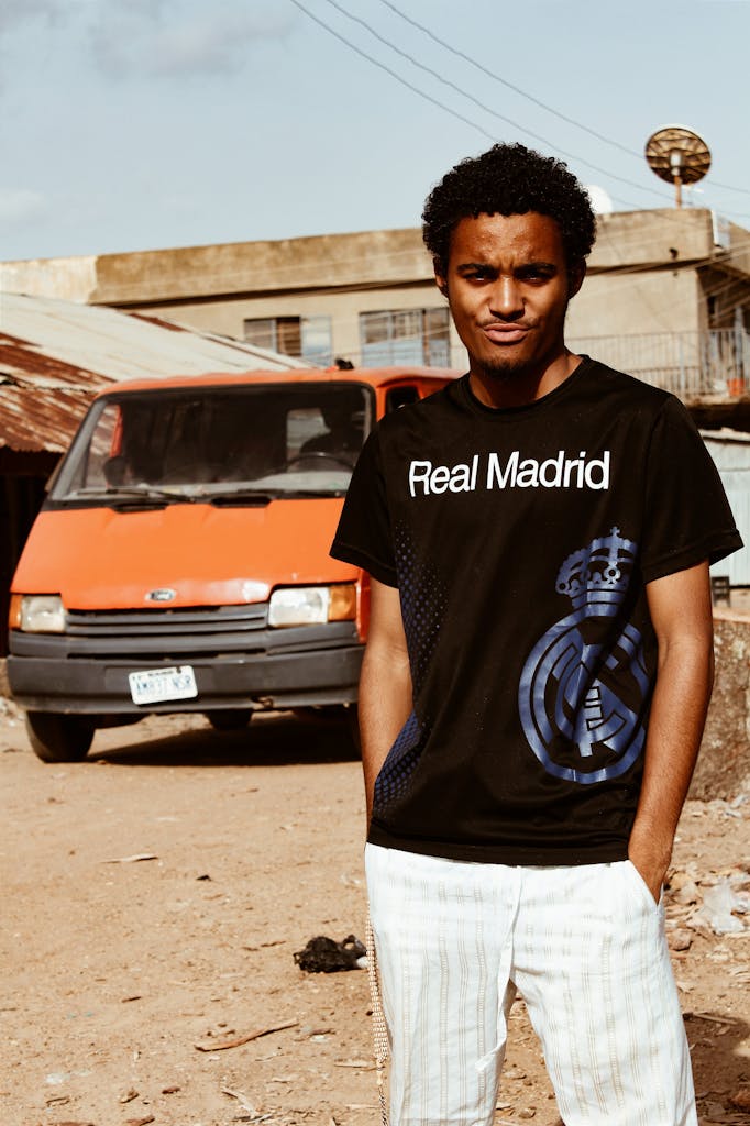 A young man poses outdoors in casual attire beside an orange van.